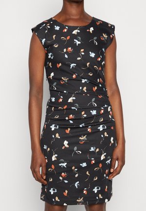 Femme portant une robe noire mi-longue à manches courtes et à motif floral multicolore, debout devant un fond uni.