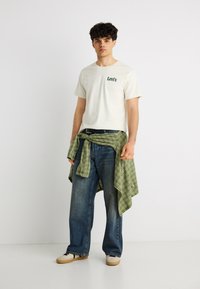 Νεαρός άνδρας που φοράει λευκό t-shirt Levi's, μπλε τζιν, μπεζ αθλητικά παπούτσια και πράσινο πουκάμισο καρό δεμένο γύρω από τη μέση του, σε λιτό φόντο.