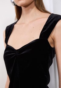 Vestido de terciopelo negro con escote cuadrado, tirantes anchos y detalle fruncido en el centro. Textura suave y silueta estructurada.