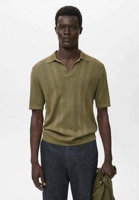 Olivgrünes Kurzarm-Polo-Shirt mit vertikaler Rippstruktur und V-Ausschnitt, kombiniert mit dunklen Hosen und einer leichten Jacke, die in der Hand gehalten wird.