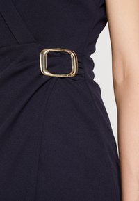 Robe bleu marine foncé avec une boucle carrée dorée serrant le tissu à la taille, présentée à côté d'un bras nu sur un fond clair.