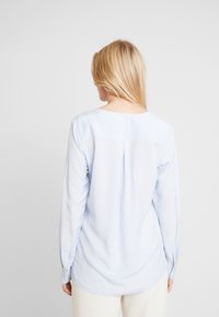 Camicia a maniche lunghe di colore blu chiaro, tessuto liscio, orlo leggermente curvo, dettaglio di piega sul retro, pattina con bottoni nascosta, con polsini con bottone.