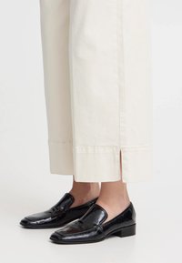 Mocassini neri in vernice con punta quadrata e tacco basso, abbinati a pantaloni larghi color crema con spacchi laterali all'orlo.