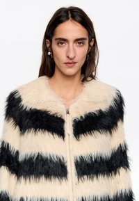 Nep-vachtjack dat is voorzien van afwisselend zwarte en crèmekleurige strepen, een brede kraag en een ritssluiting aan de voorkant. Hoogpolige textuur met een zachte uitstraling.