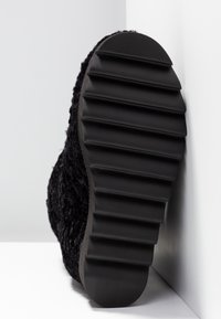 Suela negra con profundas estrías en ángulo contra una esquina blanca, unida a una parte superior de zapato de tela oscura y texturizada.