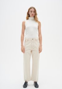 Vit ribbad ärmlös topp med hög krage; beige raka jeans med synliga sömmar och framfickor; svarta snörskor.