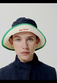 Jeune personne portant un bob bleu marine et blanc avec une bordure verte et l'inscription "Bobo Choses" en rouge, associé à une veste sombre à col montant.