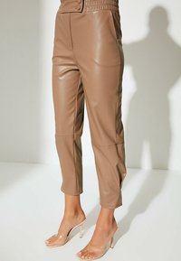 Braune Fake-Leder-Cropped-Hosen mit einem gerafften Bund, Seitennähten und glatter Textur, kombiniert mit transparenten Riemchen-Absätzen.