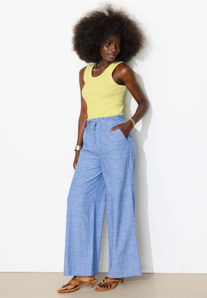 Femme aux grands cheveux bouclés portant un haut sans manches jaune, un pantalon large bleu rayé et des sandales marron à talons, debout contre un mur blanc.