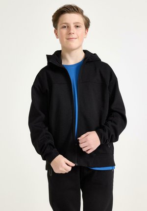 Teenagedreng med kort lysebrunt hår iført en sort hættetrøje med lynlås over en blå skjorte, stående og lynende lynlåsen.