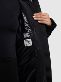 Fekete kabát sima belső béléssel. Címkék szöveggel. A szövet melegnek tűnik, plüssös textúrával a gallér környékén.