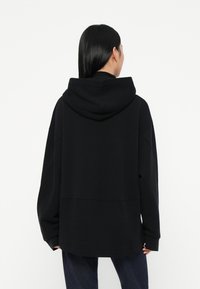 Crna hoodie s opuštenim krojem, s velikom kapuljačom i dugim rukavima. Glatka tekstura, istaknuti detalji šavova i jednostavan dizajn.