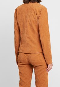 Tvådelat outfit i mjukt, texturerat tyg i rostfärg. Anpassad kavaj med enkel krage, i kombination med matchande smalbyxor.