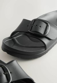 Schwarze Slides-Sandalen mit strukturierten breiten Riemen und großen quadratischen Schnallen auf einer hellen Oberfläche.