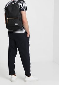 Zwarte rugzak met een voorvak met rits, een beige ritstrekker en een Herschel-label. Draagt donkerblauwe broek en witte sneakers. Gestreept hemd zichtbaar.