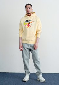 Moletom amarelo da Nike com estampa gráfica e bolso tipo canguru, combinado com calças de moletom cinzas e sapatilhas brancas, em pé contra um fundo simples.