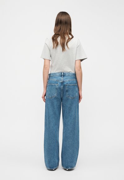 Άτομο με μακριά καστανά μαλλιά, φοράει γκρι κοντομάνικο πουκάμισο και μπλε τζιν παντελόνι φαρδιάς γραμμής, στέκεται γυρισμένο με την πλάτη προς τα εμπρός, σε απλό λευκό φόντο.