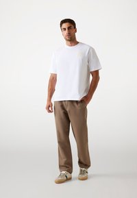 Hvid bomulds T-shirt med korte ærmer og et firkantet logo-patch, parret med brune chinos og beige sneakers med grønne detaljer.