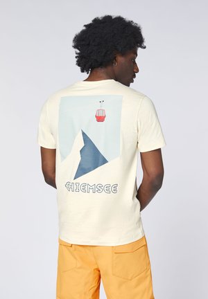 Man with curly hair wearing a cream T-shirt with a mountain and red cable car graphic and the word "Chiemsee" on the back, and orange shorts.