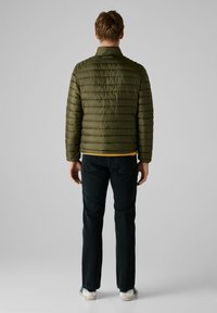 Chaqueta acolchada de color verde oliva con un diseño de costura, cuello y un dobladillo amarillo. Combinada con jeans oscuros y zapatos de suela blanca.