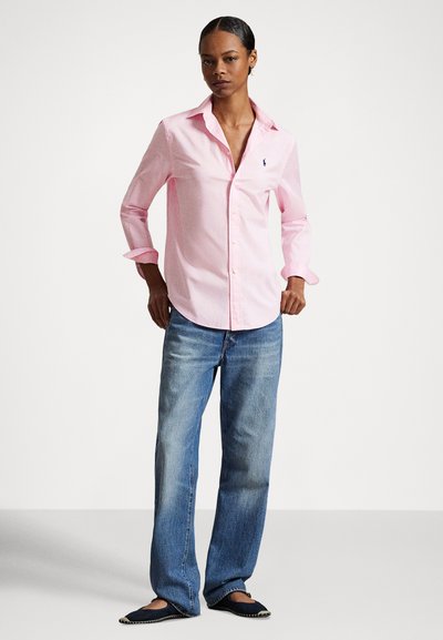 Moteris stovi rankomis ant klubų, dėvinti šviesiai rožinę mygtukais užsegamą palaidinę, mėlynas džinsus ir juodus plokščius batus, fone – paprastas baltas fonas.