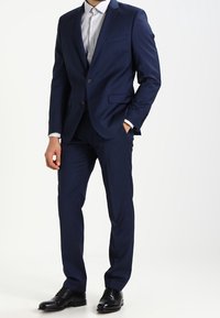 Costume bleu marine à coupe ajustée, comprenant une veste à un boutonnage, un pantalon à plis plats et une chemise de couleur claire en dessous.
