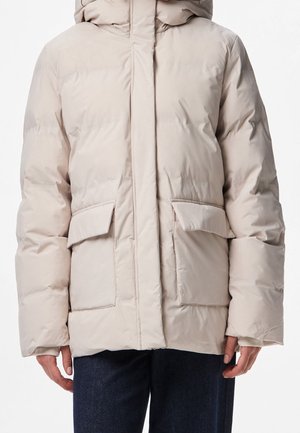 Personne portant une veste matelassée beige clair avec de grandes poches avant et une capuche, associée à un jean bleu foncé.