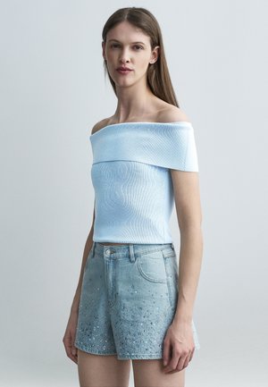 Femme portant un haut bleu clair à épaules dénudées et un short en jean taille haute avec de petites décorations, debout devant un fond uni.