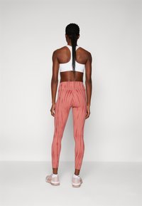 Mulher com cabelo trançado, vestindo um top desportivo branco, leggings rosas com padrão e ténis rosa claro, de pé voltada para uma parede branca.