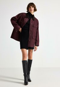 Giacca oversize borgogna con colletto marrone, abbinata a un vestito nero e stivali neri sopra il ginocchio, esposta in un contesto minimalista.
