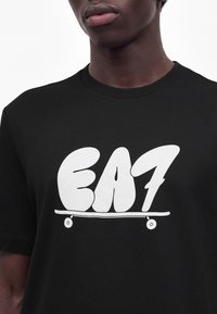 Μαύρη μπλούζα με μεγάλα λευκά γράμματα "EA7" πάνω από ένα απλό σκίτσο σκέιτμπορντ στο στήθος, φορεμένη από άτομο με σκούρο δέρμα.
