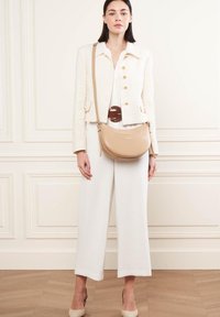 Sac à épaule en cuir beige avec une finition lisse, une forme courbée et un logo discret. Le mannequin porte une combinaison blanche et une veste crème texturée.