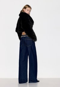 Blonde Frau in dunklen, weit geschnittenen Jeans und schwarzer, flauschiger, kürzer geschnittener Jacke, steht mit dem Rücken zur Kamera, den Kopf zur Seite gedreht, vor einem schlichten weißen Hintergrund.