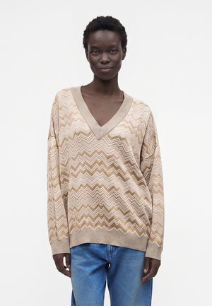 Femme aux cheveux courts et bouclés portant un pull à col en V beige et blanc à motif zigzag et un jean bleu, debout devant un fond blanc uni.