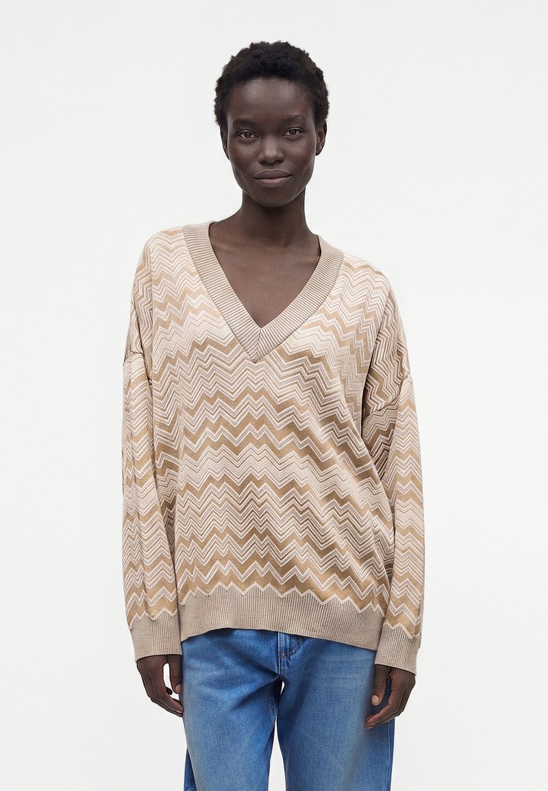 Femme aux cheveux courts et bouclés portant un pull à col en V beige et blanc à motif zigzag et un jean bleu, debout devant un fond blanc uni.