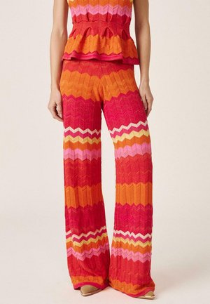 Mujer con pantalones de punto de talle alto y pierna ancha con rayas en zigzag en tonos rojo, naranja, rosa, amarillo y blanco, combinados con una camiseta a juego.