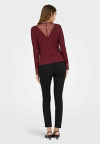 Bordeaux kleurig langsliefde shirt met een kanten V-achterkant en bijpassende kanten manchetten, aansluitend ontwerp, gecombineerd met zwarte skinny jeans en zwarte hoge hakken.