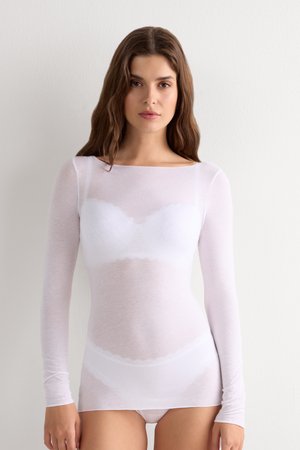Femme portant un haut transparent blanc à manches longues par-dessus un soutien-gorge et une culotte blancs, debout contre un mur clair uni, face à la caméra.