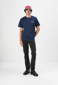 Camiseta azul marino de manga corta con un pequeño detalle rojo bordado en el pecho, combinada con unos vaqueros negros y zapatos negros. Lleva una gorra rosa.