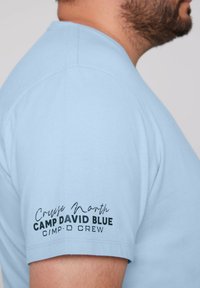 Camiseta de manga corta azul claro con texto negro "Cruise North CAMP DAVID BLUE CMP-D CREW" en la parte superior del brazo izquierdo. Textura de tela suave.