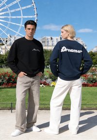 Un homme et une femme se tiennent dehors près d'une grande roue ; l'homme porte un sweat noir avec l'inscription « Miro Paris », la femme porte un sweat bleu marine avec « Dreamer » dans le dos.