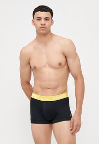 Črne bokserice z rumenim logotipom Nike na pasu, ki nudijo tesen oprijem in gladko teksturo. Model stoji z rokama za hrbtom.