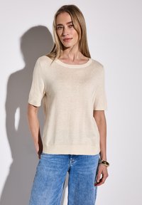 Top corto lavorato a maglia di colore crema con scollatura rotonda e vestibilità rilassata, abbinato a jeans a gamba larga di colore blu chiaro. Texture liscia, dettagli minimi.