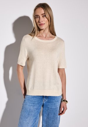 Top corto lavorato a maglia di colore crema con scollatura rotonda e vestibilità rilassata, abbinato a jeans a gamba larga di colore blu chiaro. Texture liscia, dettagli minimi.