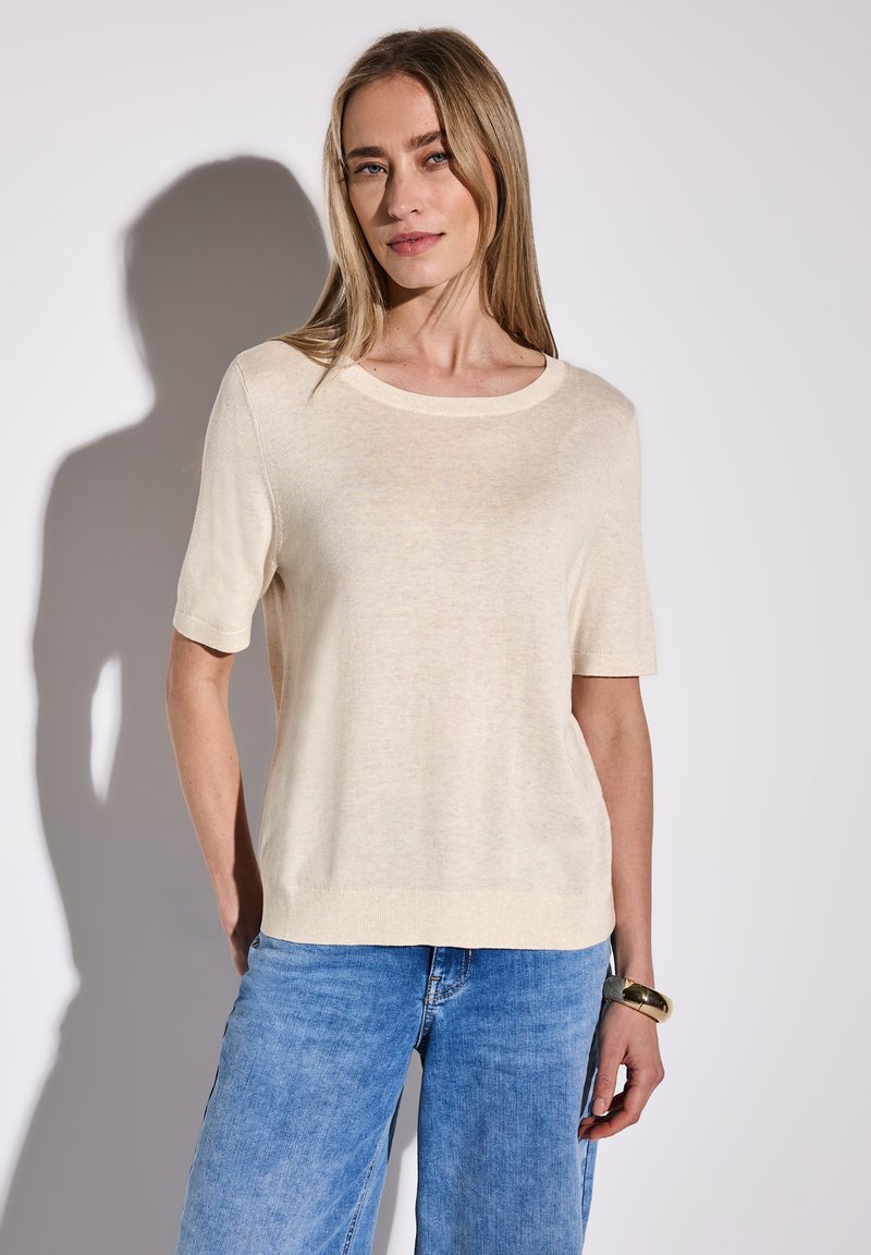 Top corto lavorato a maglia di colore crema con scollatura rotonda e vestibilità rilassata, abbinato a jeans a gamba larga di colore blu chiaro. Texture liscia, dettagli minimi.