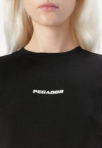 T-shirt preta de algodão com um logótipo branco bordado "PEGADOR" no peito. O tecido parece liso, com um design clássico de gola redonda.