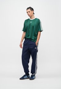 Mladić s pletenicama, obučen u zelenu Adidas majicu, tamnoplave Adidas trenirke i plave Adidas tenisice, stoji s rukama u džepovima.