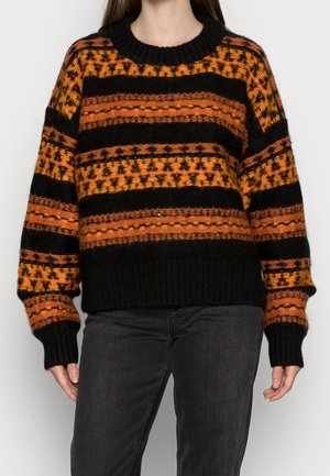 Femme portant un pull en maille noir et orange à motifs avec des manches longues et un pantalon noir sur un fond uni.