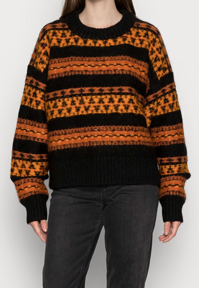 Femme portant un pull en maille noir et orange à motifs avec des manches longues et un pantalon noir sur un fond uni.