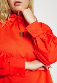 Červená mikina Nike s kapucí a stahovací šňůrkou u límce s černým vyšitým logem. Má propracované rukávy se zlatými detaily ve tvaru smyčky na manžetách.